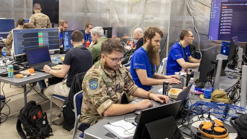 10 personer sitter vid skrivbord i en lokal som är fylld med datorer, skärmar, sladdar och elektronisk utrustning. Några av dem är klädda i militäruniformer. 
