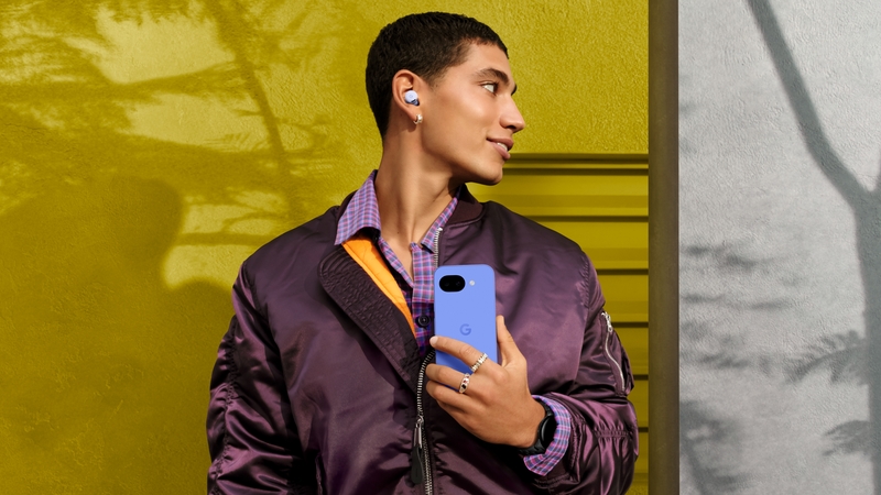 En ung man står vid en gulgrön husvägg. Han är klädd i lila jacka, har hörlurar i öronen och håller i en lila smartphone av märke Google Pixel 10a. 