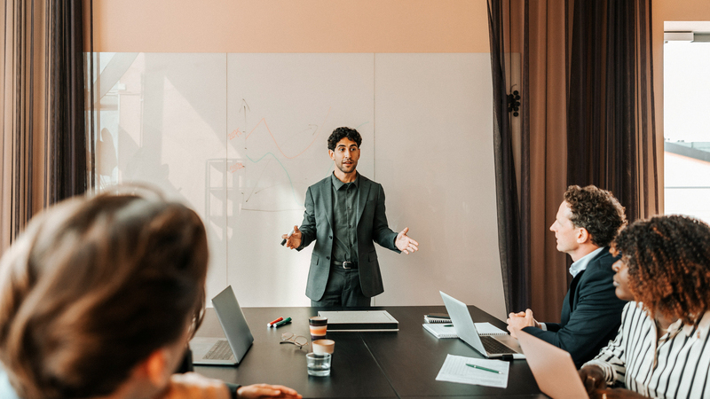 Två män och en kvinna sitter vid ett konferensbord och lyssnar till en man som står framför en whiteboard och pratar. 