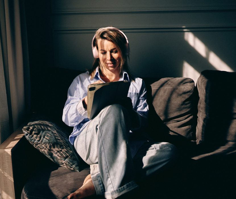 Person med hörlurar sitter avslappnat i en soffa och tittar på en surfplatta, klädd i blå skjorta och ljusa byxor.