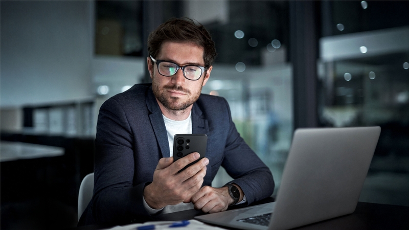 En man i kavaj sitter på en restaurang med en laptop och en mobil. 