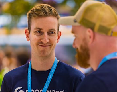 A smiling man wearing a dark blue shirt and event lanyard looks at another bearded man during a conversation in a bright, busy indoor setting.