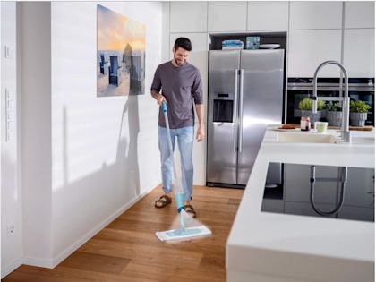 A man is mopping a wooden floor in a modern kitchen using a flat mop; in the background, a stainless steel refrigerator, a kitchen island, and plants are visible.