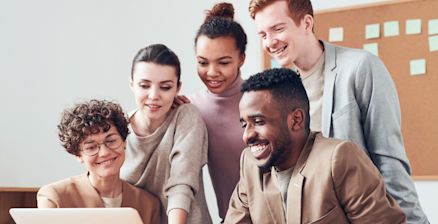 A diverse group of five young adults stands around a laptop, laughing together. They appear motivated and are collaborating as a team in a modern office with a bulletin board in the background.