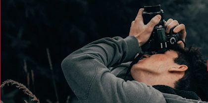 Nahaufnahme einer Person, die eine Kamera ans Auge hält und nach oben fotografiert; sie trägt einen grauen Hoodie, der Hintergrund ist dunkel und unscharf.