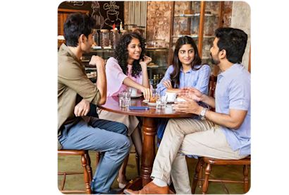 Vier junge Erwachsene sitzen gemeinsam in einem Café an einem runden Holztisch, unterhalten sich und trinken Kaffee.