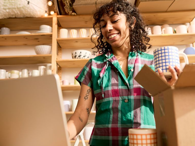 Eine Frau hält eine Tasse Tee in der Hand, während sie in ihrem Laden an ihrem Laptop arbeitet. Vor ihr steht eine offene Box, und hinter ihr sind Keramikartikel zu sehen.