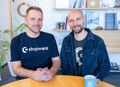 Een foto van de gebroeders Hamann (Stefan Hamann links, Sebastian Hamann rechts) in een licht kantoor. Beiden glimlachen en dragen kleding met het Shopware-logo.