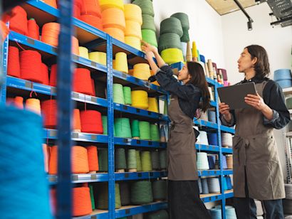 Zwei Frauen in Overalls füllen Garnspulen in einer Fabrikumgebung nach. Eine von ihnen hält ein Klemmbrett in der Hand.