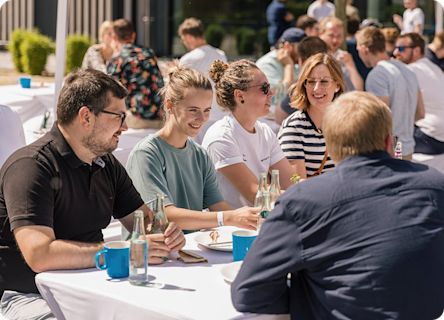 People sitting together at outdoor tables during a sunny team event, talking and enjoying food and drinks.