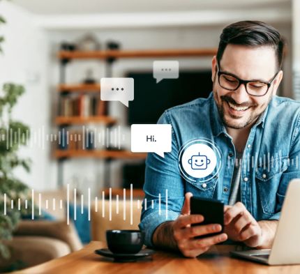 A smiling man with glasses and a beard sits at a table, using a smartphone while working on a laptop. Speech bubbles with a chatbot icon and the text "Hi." appear around him, alongside visual sound waveforms representing a conversation.