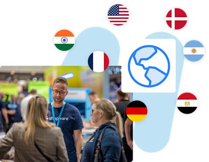 An image showcasing an international event. At the bottom, a person wearing a Shopware t-shirt is talking to two attendees in a expo setting. Above, a globe icon is displayed, surrounded by circular flags representing various countries.