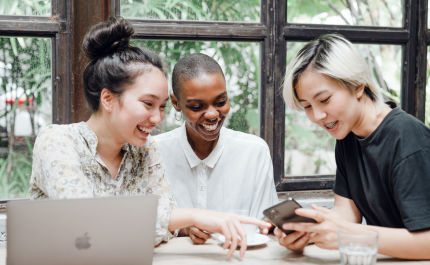 Three smiling people at a table, one holding a smartphone, laptop on the table. Scene shows joy and cooperation.