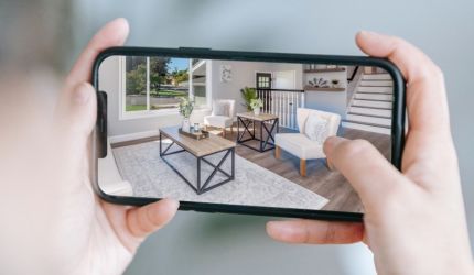 A person holds a smartphone on which a living room with furniture such as tables and armchairs is displayed in an augmented reality view, virtually furnishing the room.