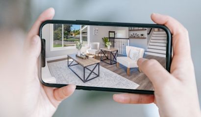 spatial commerce_1480px Eine Person hält ein Smartphone, auf dem ein Wohnzimmer mit Möbeln wie Tischen und Sesseln in einer Augmented-Reality-Ansicht dargestellt wird, wodurch der Raum virtuell eingerichtet wird.