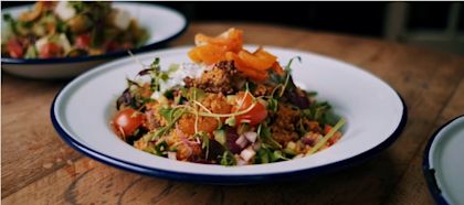 Nahaufnahme eines angerichteten Salats mit Couscous, Kirschtomaten, roten Zwiebeln, Gurken, frischen Kräutern und einem Topping aus Lachs auf einem weißen Teller mit blauem Rand; im Hintergrund ist ein weiterer Teller unscharf zu sehen.