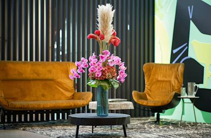 Floral arrangement in a vase on a table in a modern living room, surrounded by two orange armchairs and decorative wall design.