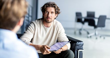 Ein Mann in einem modernen Büro hält ein Tablet in der Hand und scheint aufmerksam einer anderen Person zuzuhören, während er direkt darauf reagiert. Im Hintergrund ist ein heller Konferenzraum sichtbar, der den geschäftlichen Kontext unterstreicht.