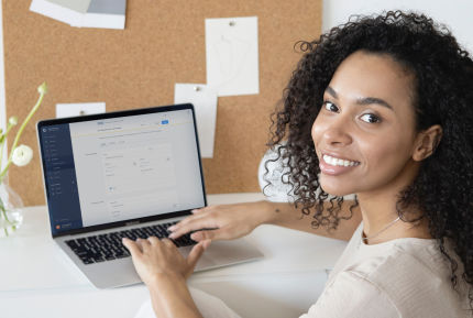 Eine Frau sitzt lächelnd vor einem Laptop und arbeitet an einer E-Mail. Im Hintergrund ist eine Pinnwand mit Notizen zu sehen. Die Szene vermittelt Produktivität und Freude an der Arbeit.