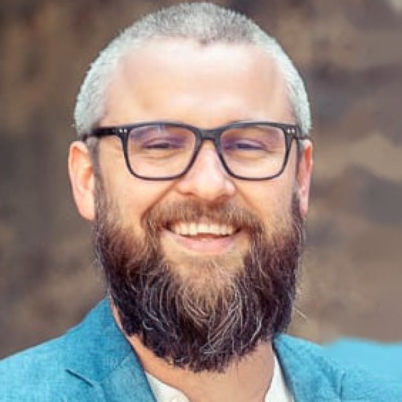 Portret van een glimlachende man met grijs haar, een volle baard en een zwarte bril.