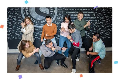 Mitarbeiter Kreidewand Konfetti Showpare-Mitarbeiter posieren spielerisch vor der Kreidewand und stellen eine Actionszene nach, wobei sie lächeln und lachen. Das Bild ist von buntem Konfetti umgeben.