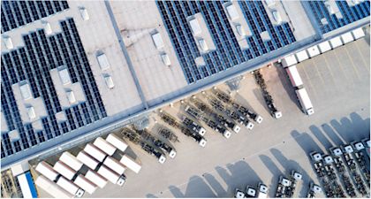 Aerial view of a large warehouse facility with solar panels covering the rooftop, loading docks along the building, and multiple trucks and trailers parked in organized rows in the distribution yard.