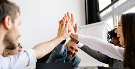 A diverse team of five people in a modern office giving a group high-five. They are laughing and appear motivated. Large windows and a flipchart are visible in the background.