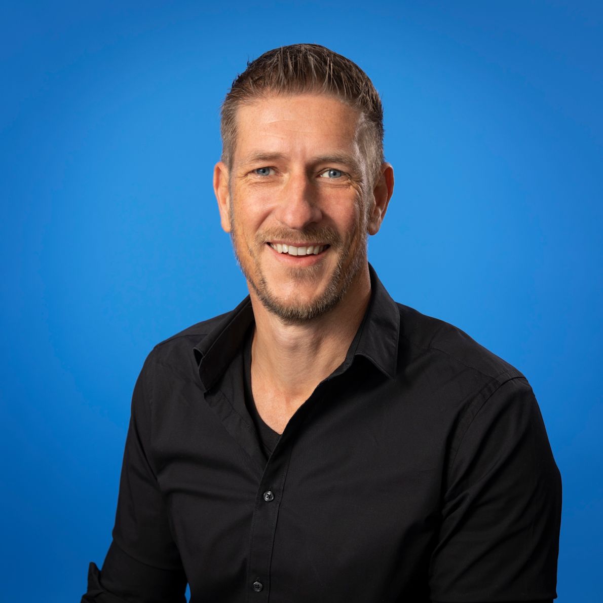 Jorrit Steinz: Portrait of a smiling man with short hair wearing a black shirt against a blue background.