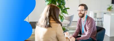 Two people are sitting and having a conversation. The man on the right is smiling, wearing glasses, and a name badge. The woman on the left, with her back to the camera, is wearing a beige coat. A plant and a modern, open office space are visible in the background.