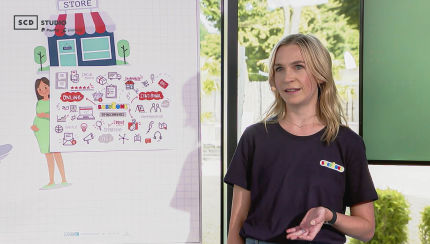 A woman is speaking and wearing a t-shirt with the BabyOne logo. Beside her is a board with various colorful icons and drawings related to online and stationary commerce, communication, and strategy.