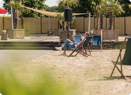 A person is reclining in a blue deck chair with legs crossed, enjoying the sunny, beach-like setting