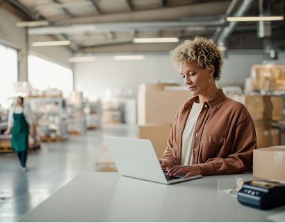 A warehouse employee uses a laptop at a packing station in a modern distribution center. Cardboard boxes and shelving are visible in the background, illustrating digital order management and warehouse operations.