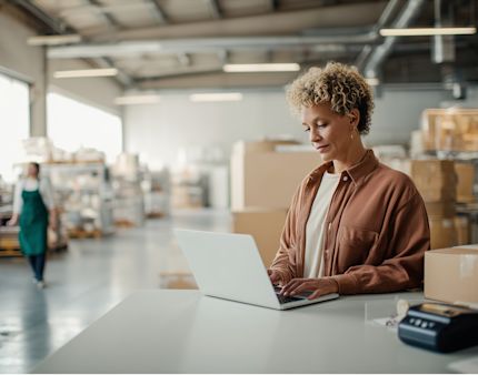 A warehouse employee uses a laptop at a packing station in a modern distribution center. Cardboard boxes and shelving are visible in the background, illustrating digital order management and warehouse operations.