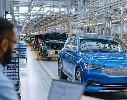 Linea di produzione automobilistica con veicoli in fase di assemblaggio, un'auto blu in primo piano e un operatore che monitora il processo su un computer portatile.