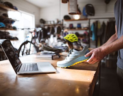 A person holds a light blue athletic shoe over a wooden counter next to an open laptop in a bicycle and outdoor gear shop.