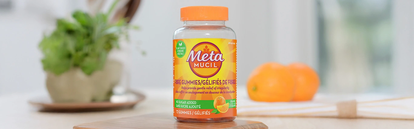 A bottle of Metamucil fibre no sugar added 72 counts placed on a wooden table, with a plate holding two oranges in the background.