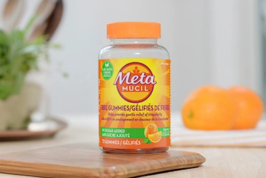 A bottle of Metamucil fibre no sugar added 72 counts placed on a wooden table, with a plate holding two oranges in the background.