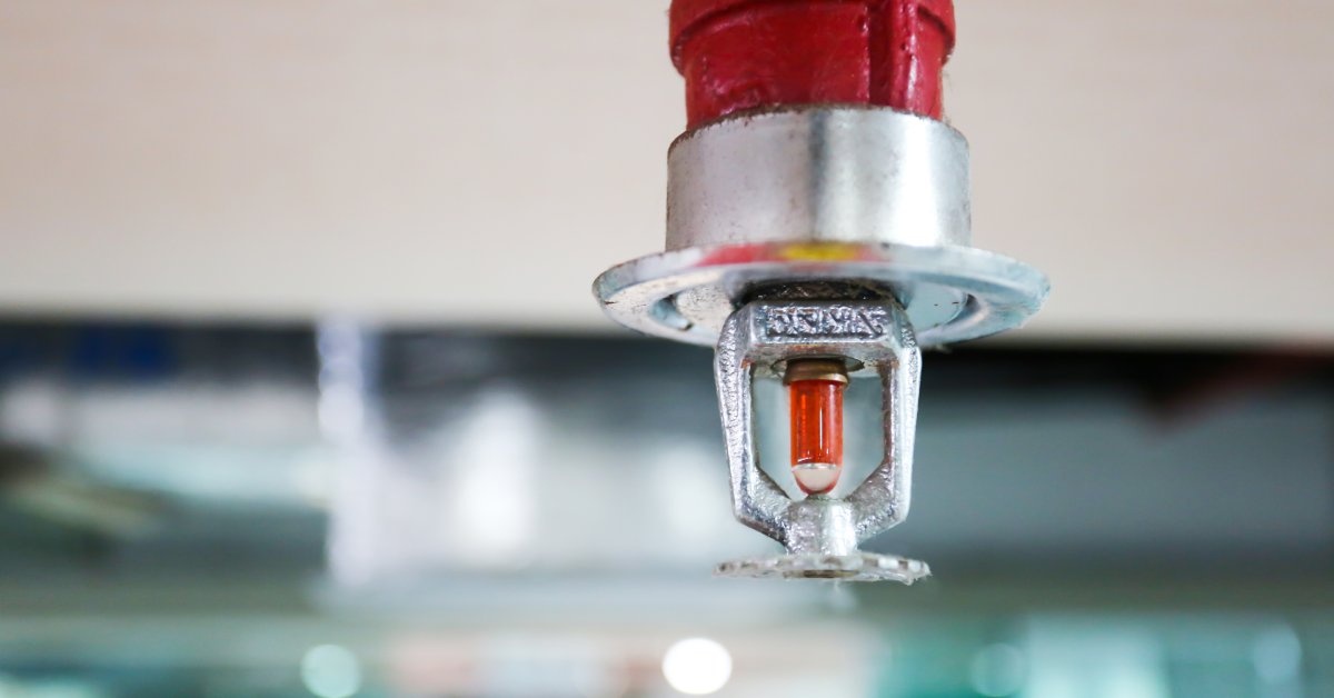 A fire sprinkler with a silver tip and a red base. A drop of water is suspended from the nozzle, and the background is blurred.