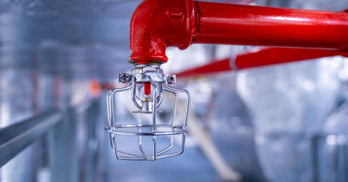 A red pipe on a wall connects to a fire sprinkler, which features a metal cage around the sprinkler head.