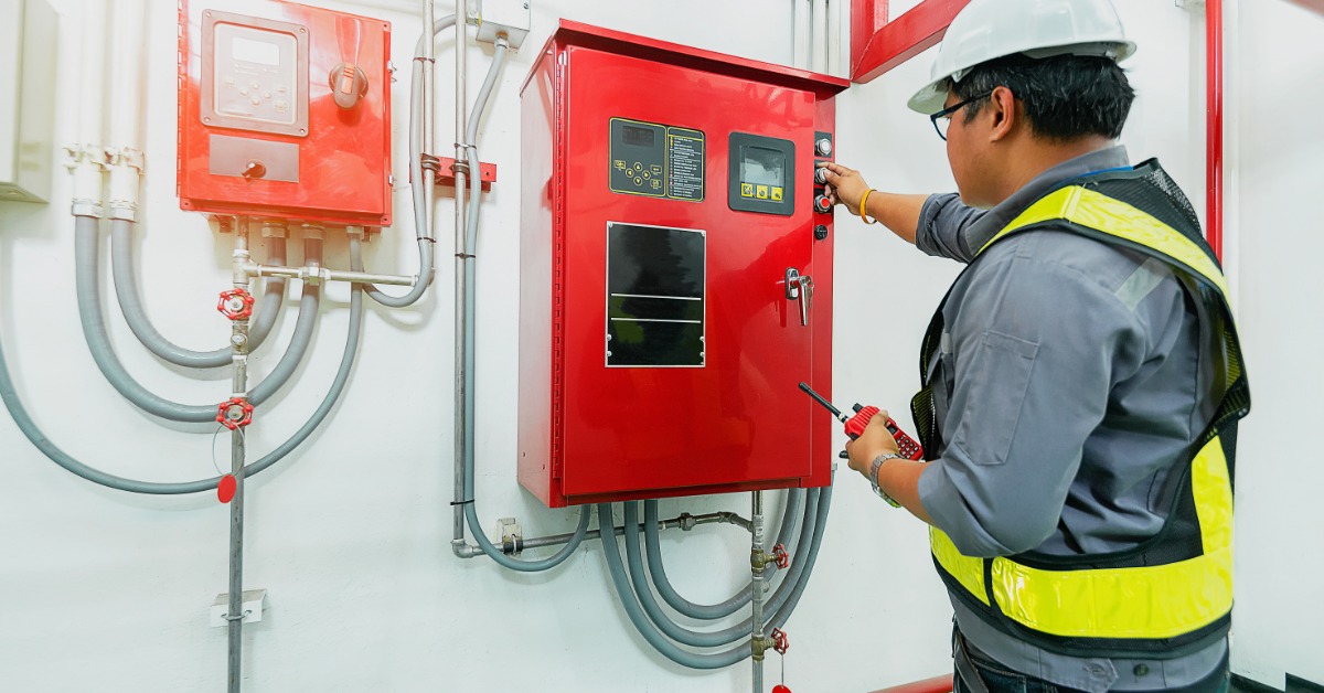 An engineer in a hard hat and a safety vest stands in front of the controls for a building's sprinkler system.