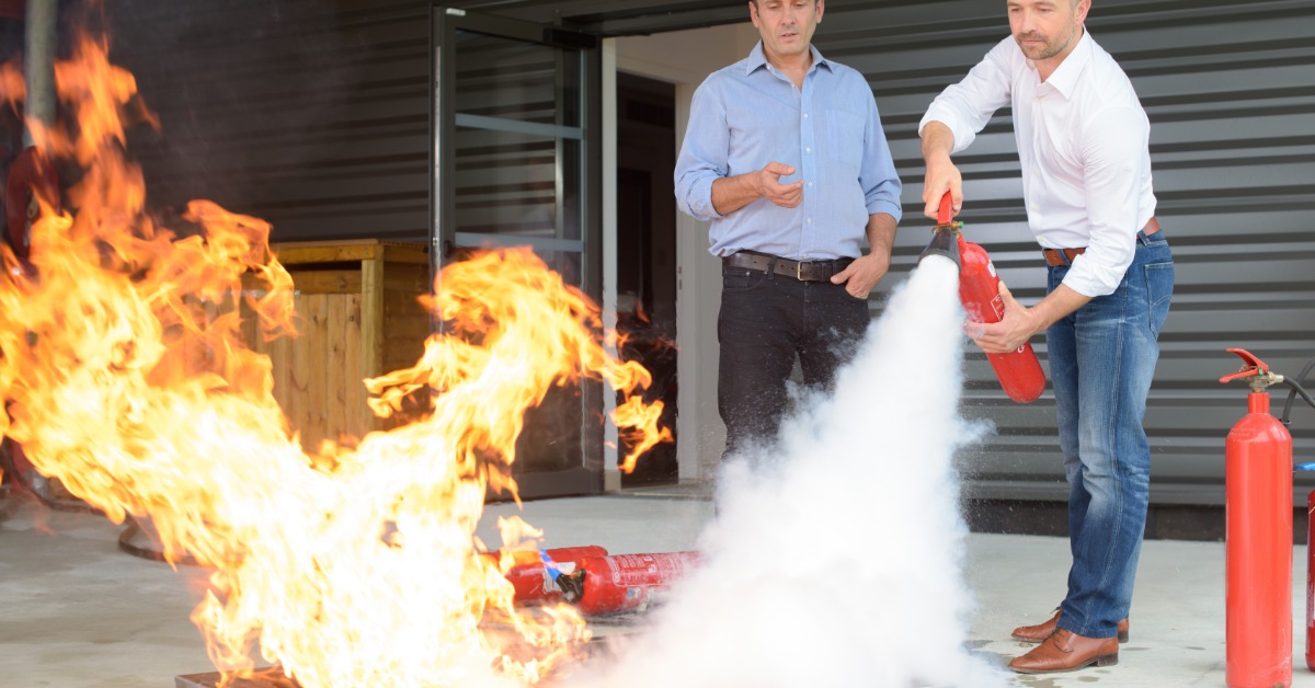 A man in a white shirt uses a red fire extinguisher to put out flames during an outdoor fire-safety training drill.