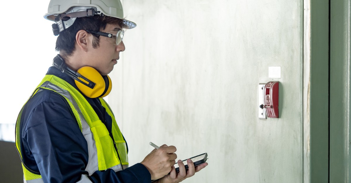 A building inspector looks at a fire alarm system on a wall and makes marks on his smartphone. He wears a hard hat.