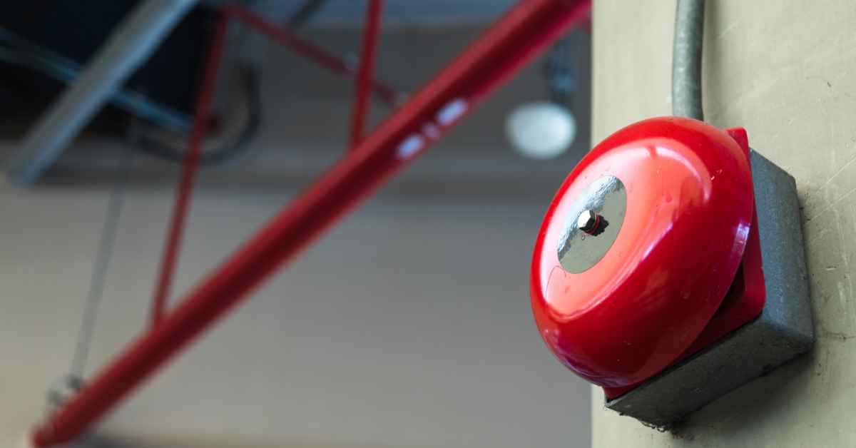 A fire alarm sits on a wall in a facility and features a large red bell. Red piping hangs in the background.
