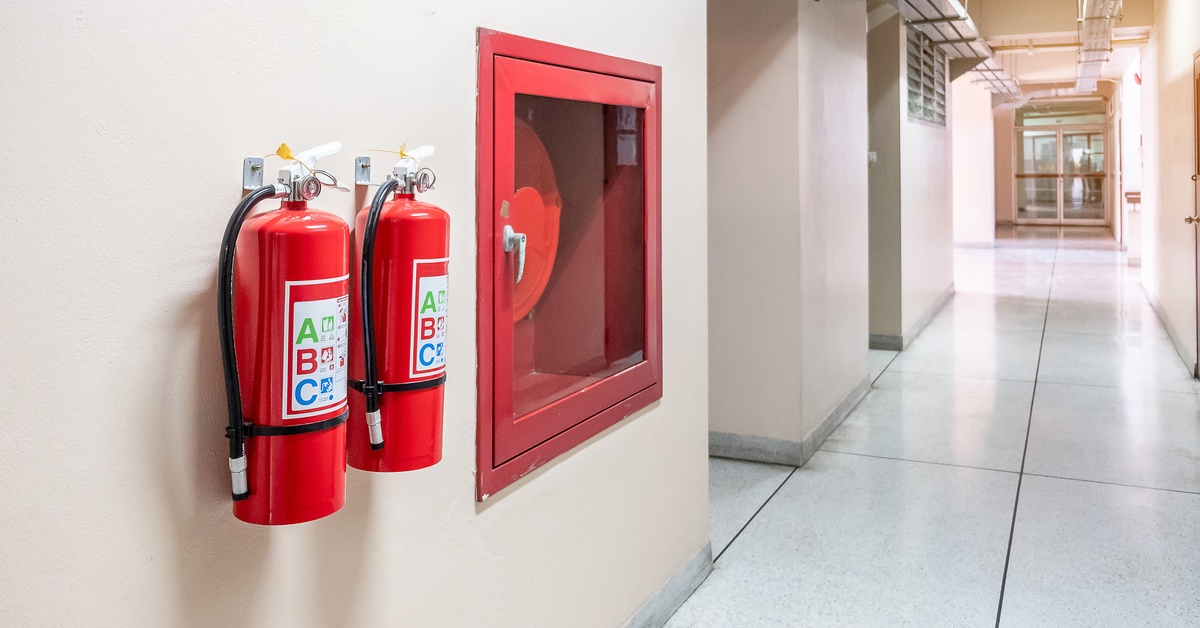 Two red fire extinguishers hang on a wall in a building hallway featuring the letters ABC on their labels.