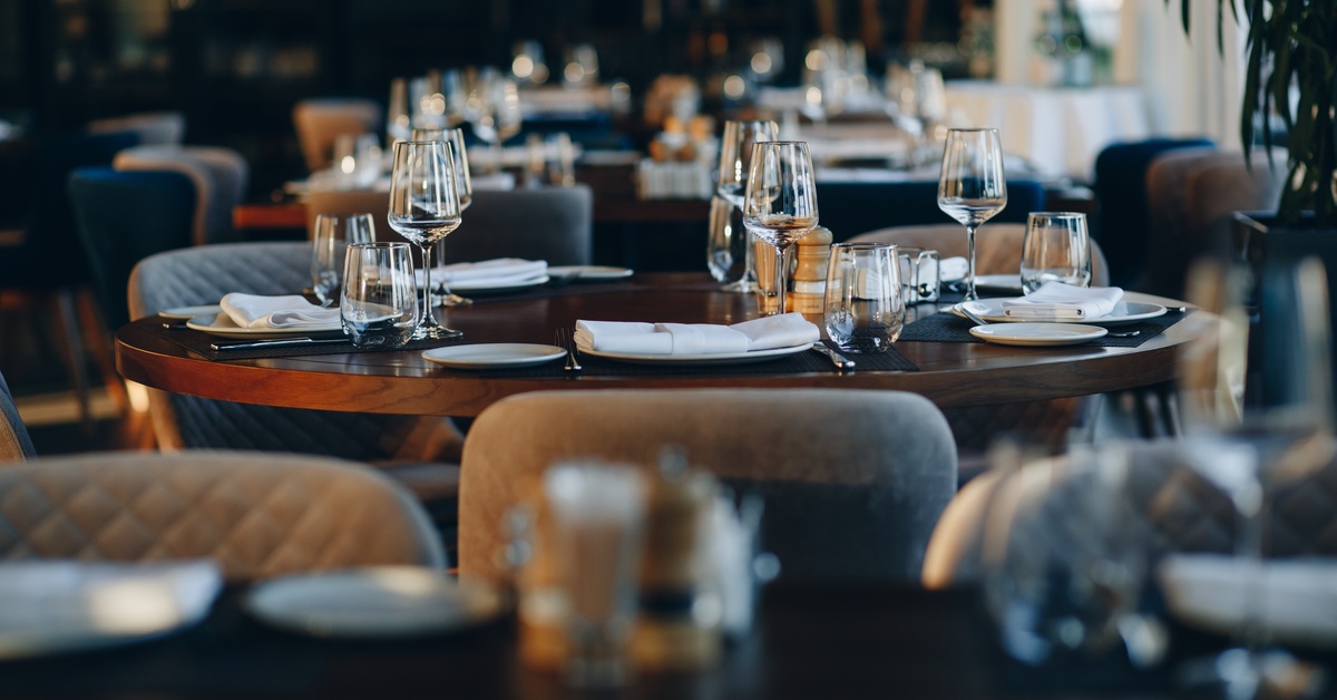 Multiple empty tables in a restaurant feature glassware, plates, and napkins. Tan chairs sit around each table.