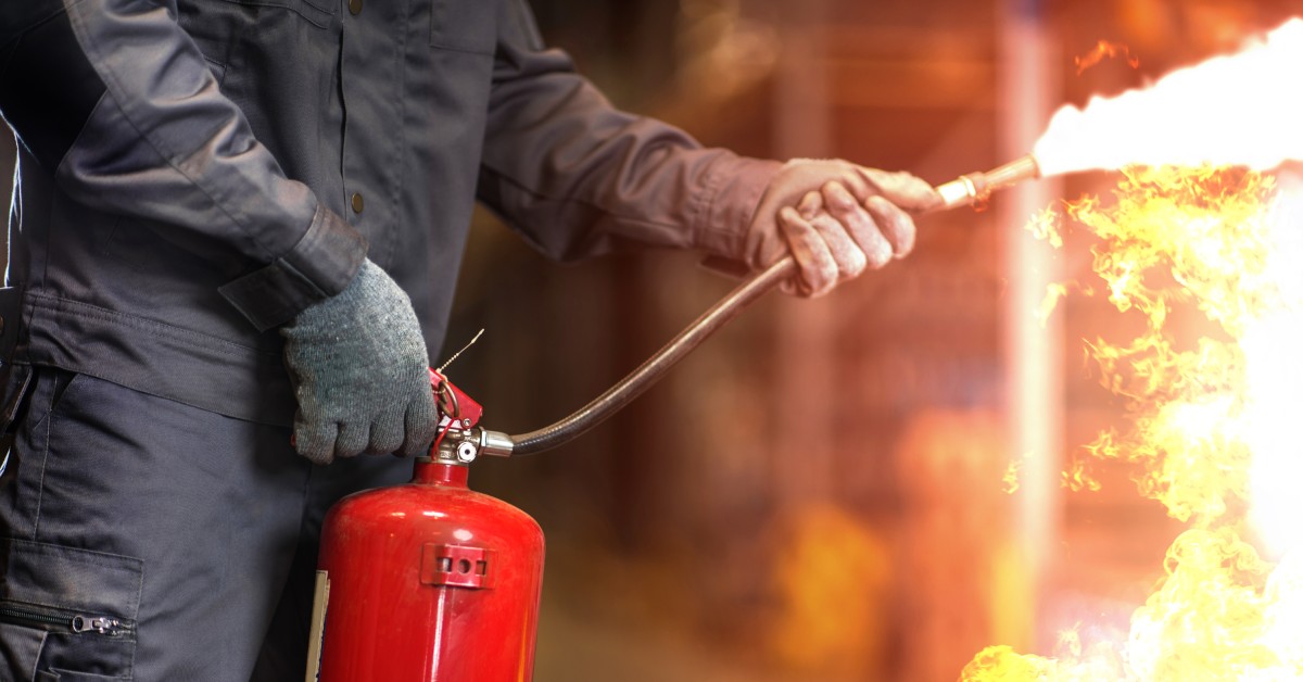 A man operates a fire extinguisher to put out a blazing fire. He wears a blue uniform and gray gloves.