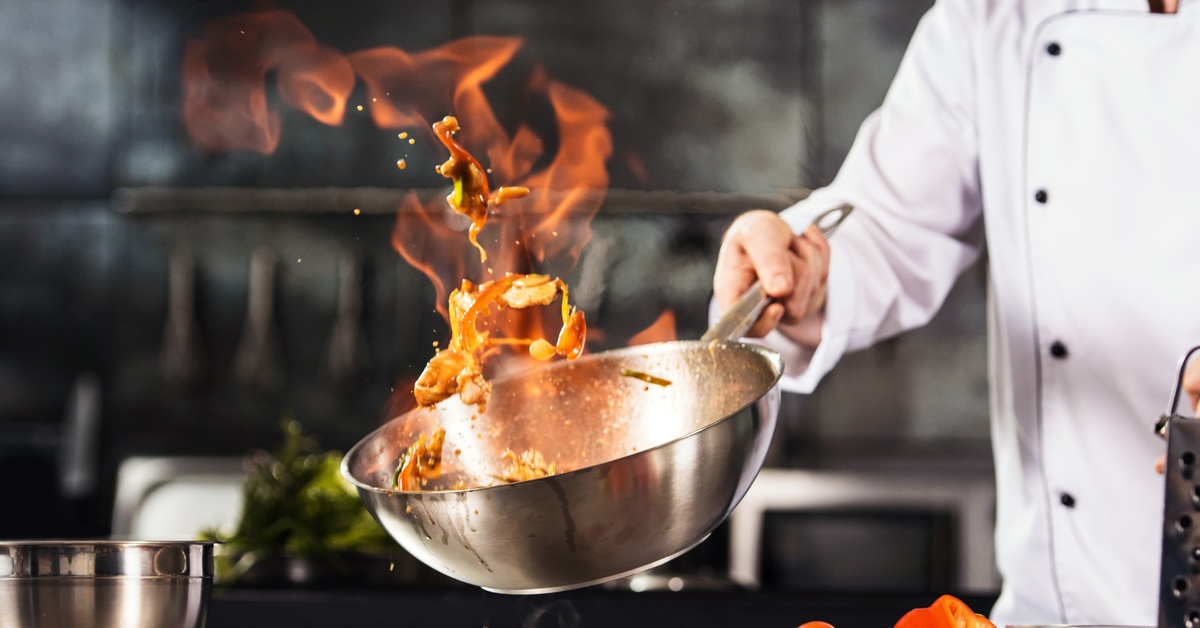 A chef holds a pan of food in a kitchen, as its contents move around a flame. Tomatoes are on the counter.