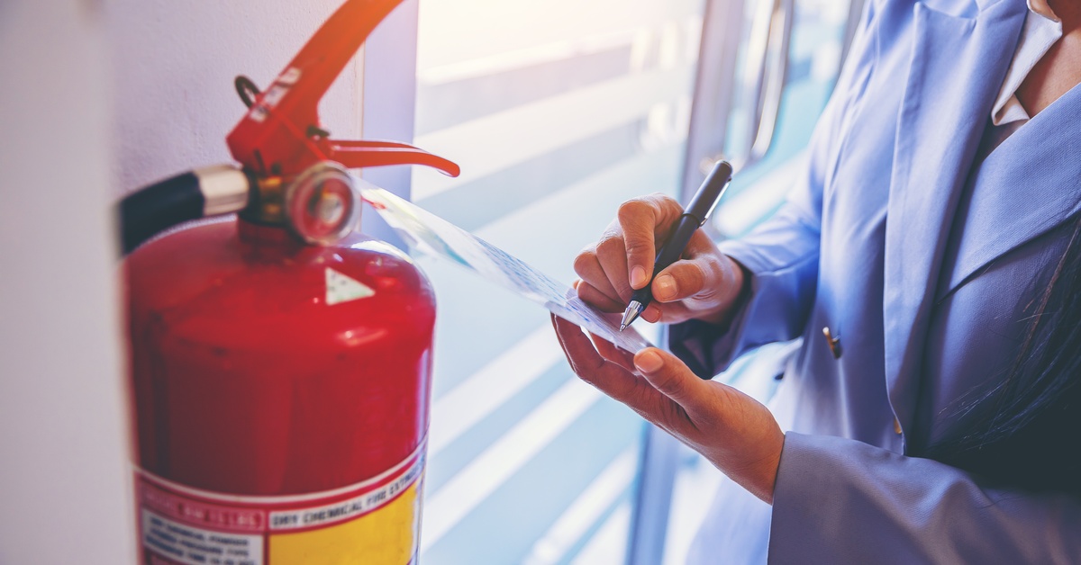 A woman makes notes on a fire extinguisher tag as she conducts an inspection. The extinguisher hangs on a wall.