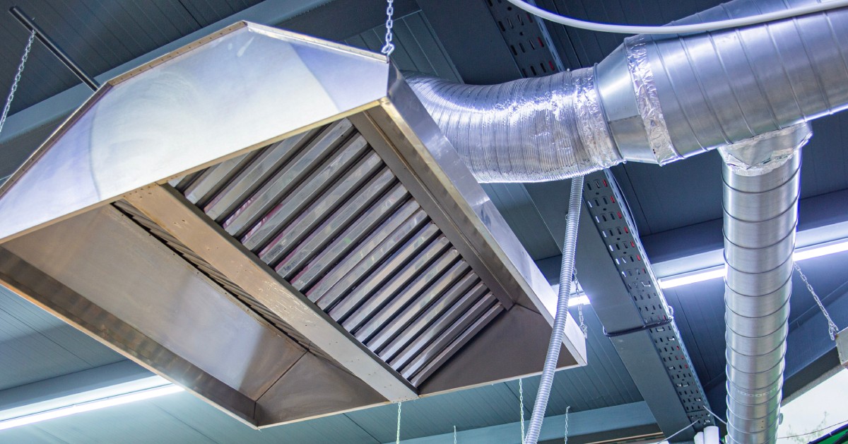 A stainless steel exhaust hood in the ceiling of a kitchen features a grease trap. It hangs next to bright lights.