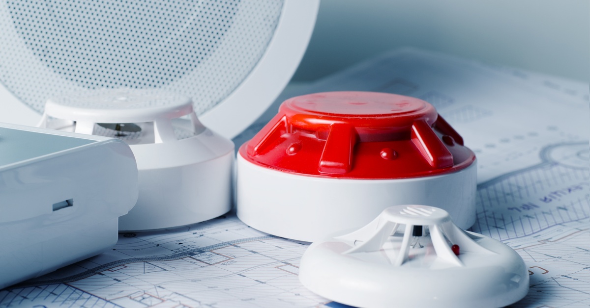 Fire alarm systems sit on rolled-out building blueprints. Most of the systems are white, and one features a red top.